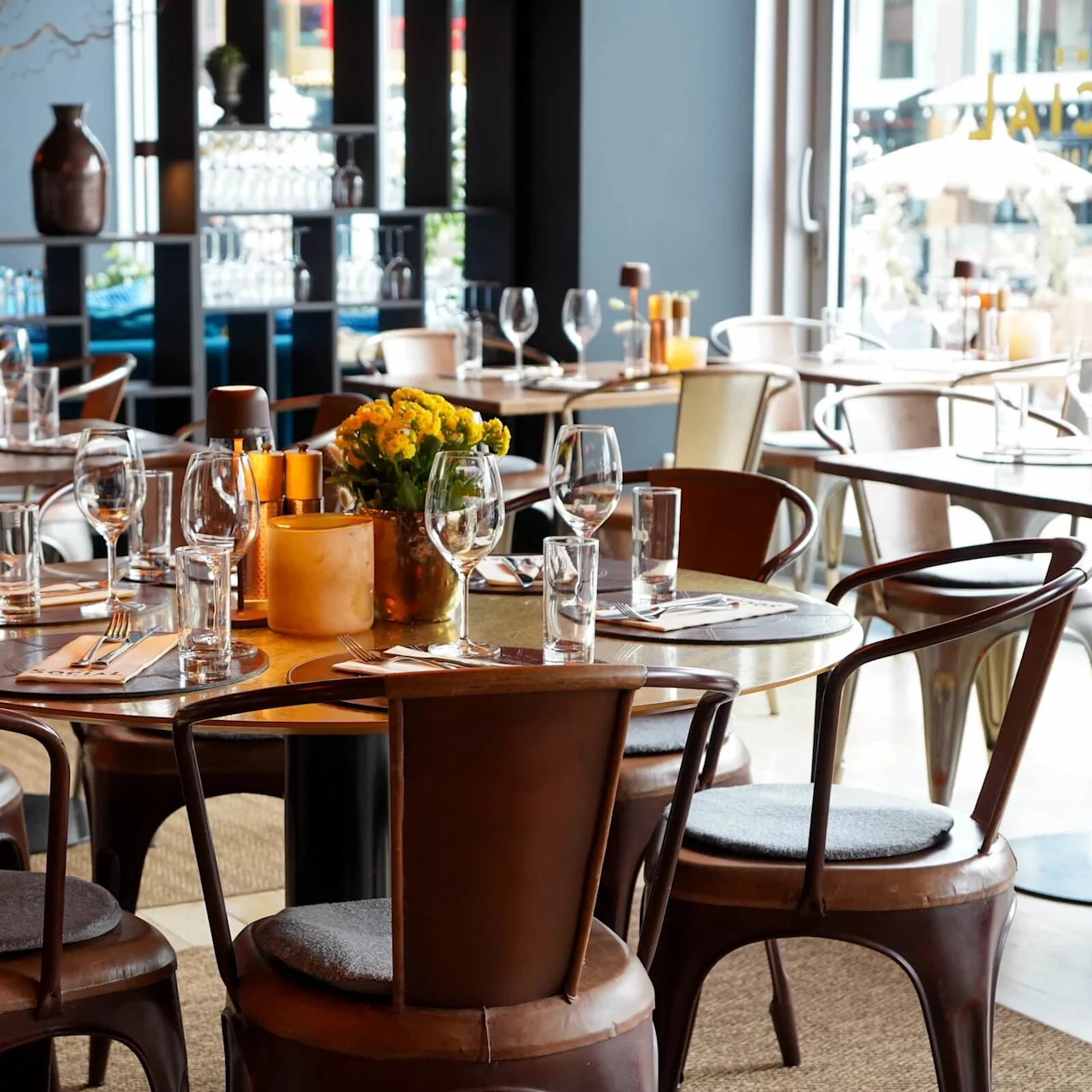 A set table with flowers and glassware, arranged on wooden chairs in a cozy, warmly lit restaurant, with large windows and decorative vases visible in the background.
