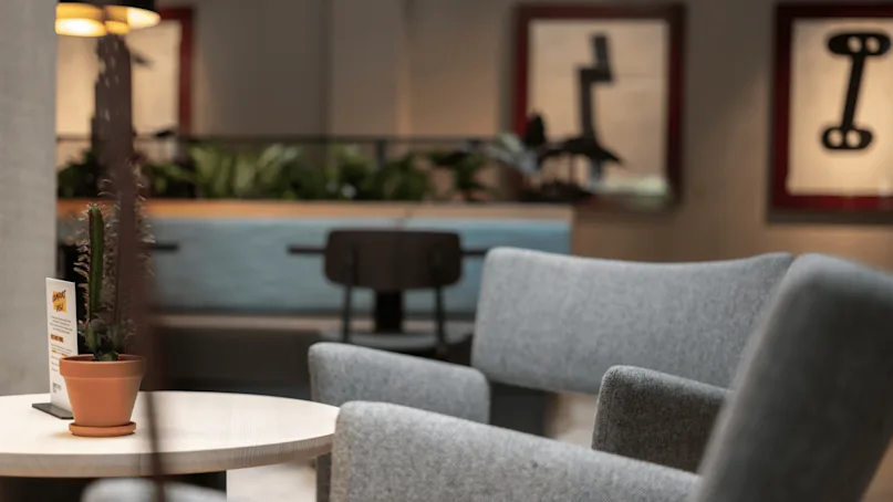 Gray armchairs surround a wooden table with a potted cactus. A sign reads "BRUNCH MENU". The background features plants, abstract art, and warm lighting.