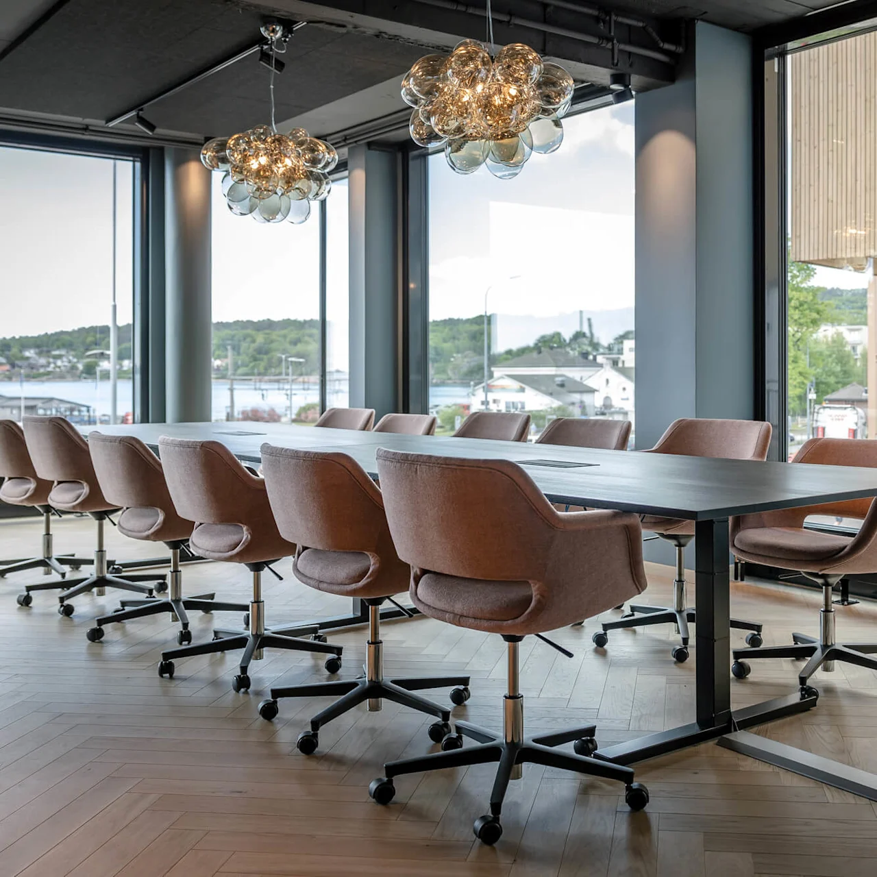 Konferencebord omgivet af drejelige stole. Store vinduer afslører en naturskøn udsigt over en sø med grønne bakker i baggrunden. Moderne pendellamper hænger ovenover.