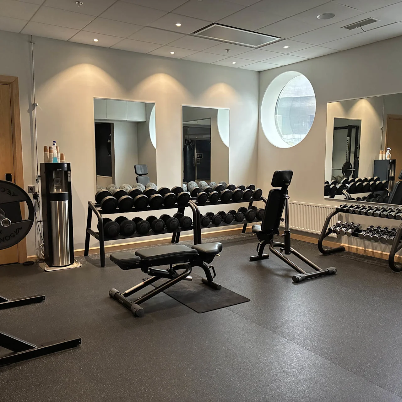 En vægtbænk står i et motionscenter, omgivet af håndvægte på stativer. Spejle reflekterer udstyret, og runde vinduer oplyser rummet. En vandkøler med kopper er i nærheden.