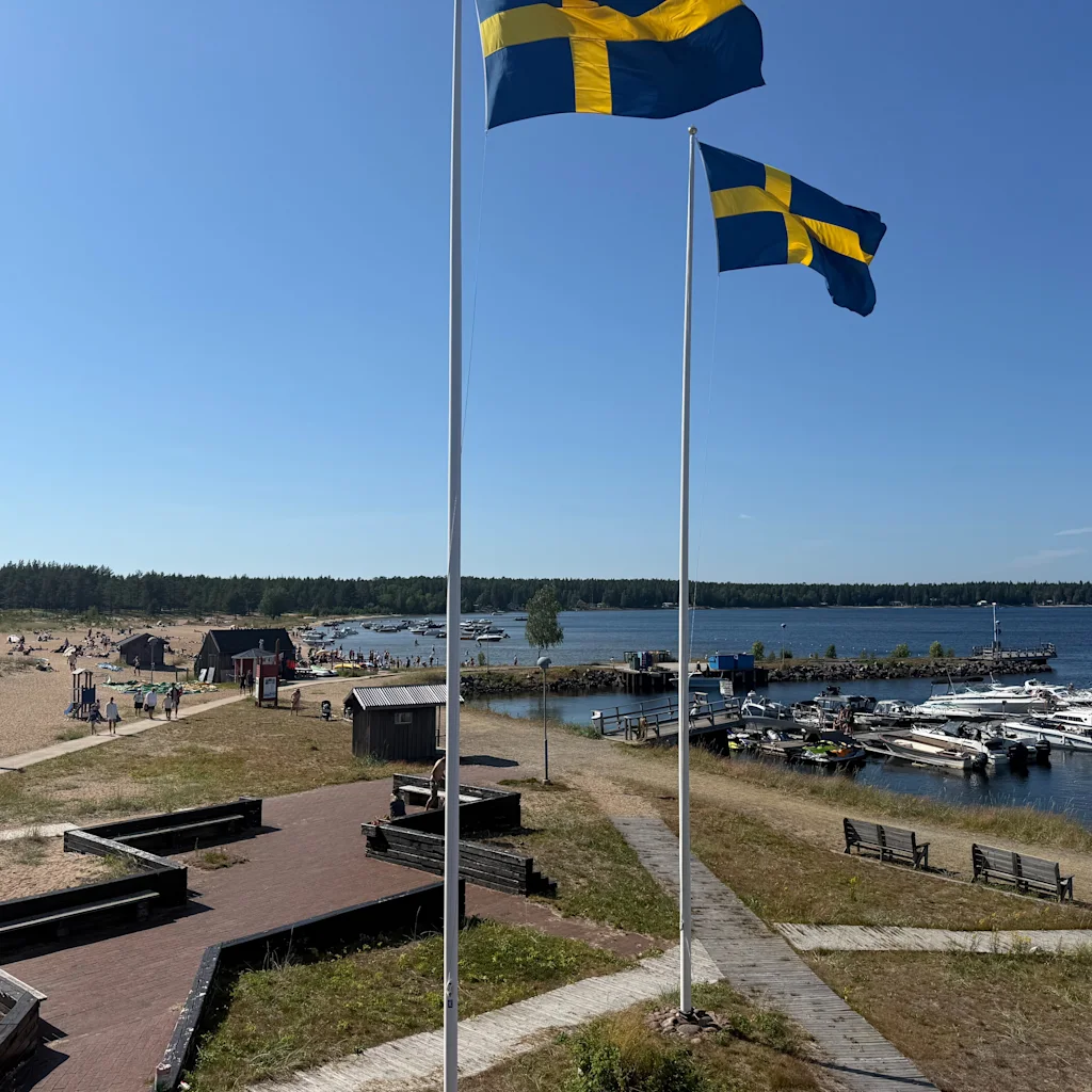 Swedish flags flying over a sunny waterfront with a beach, people, and boats docked at a marina.