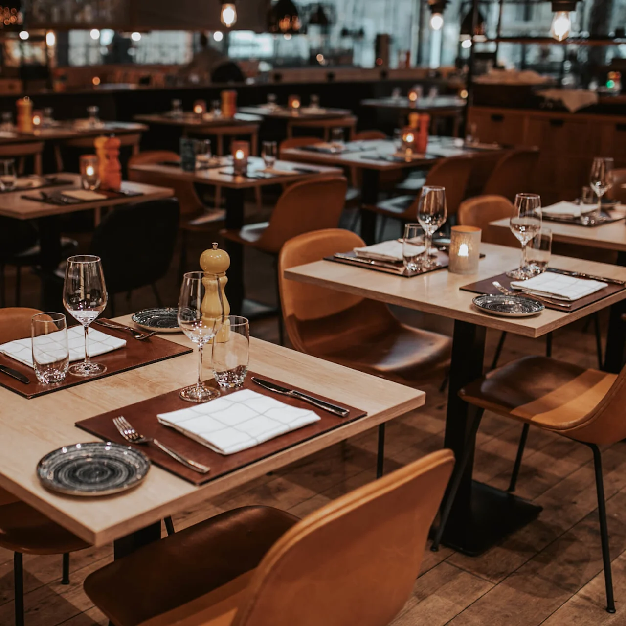 Tables set for dining, featuring wine glasses, plates, and folded napkins, create a cozy ambiance in a warmly lit restaurant with wooden flooring and neatly arranged chairs.