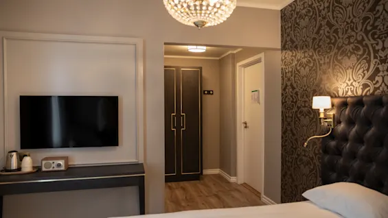 Et velindrettet hotelværelse med en seng, et TV, et klædeskab og mønstret tapet. En lysekrone og væglampe giver belysning.