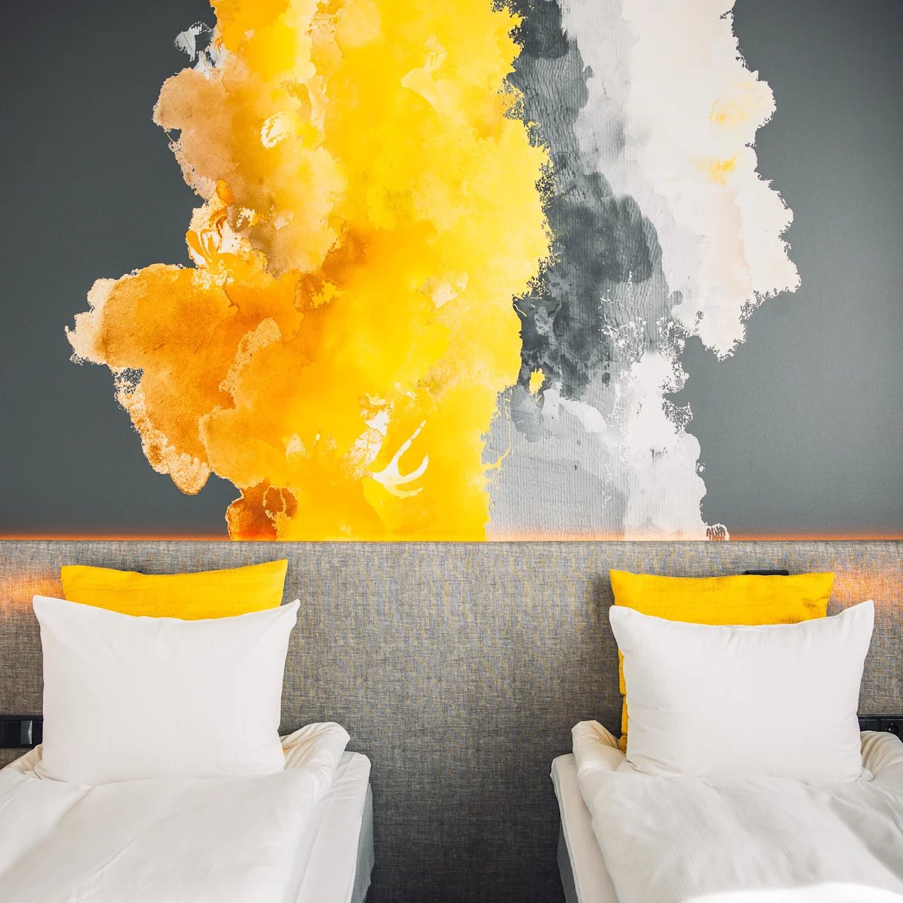 Two beds with white linens and yellow pillows against a dark wall. A large abstract art piece with splashes of yellow, gray, and white decorates the wall, adding vibrant contrast.