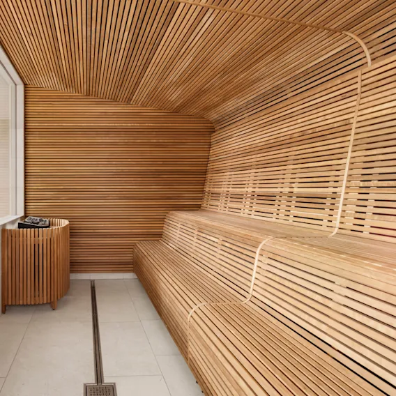 A wooden sauna features horizontal slatted benches and walls with a central heater, surrounded by a clean, modern spa area visible through a glass partition.