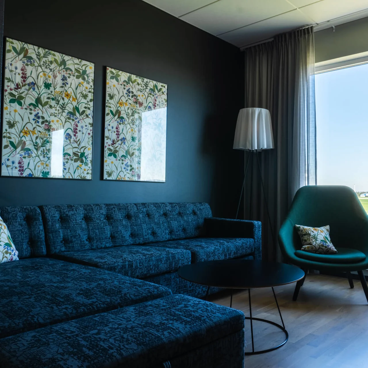 En blå sittsoffa och grön stol med blommiga kuddar står i ett vardagsrum. Två botaniska prints hänger på en mörk vägg nära ett stort fönster med utsikt över ett soligt landskap.