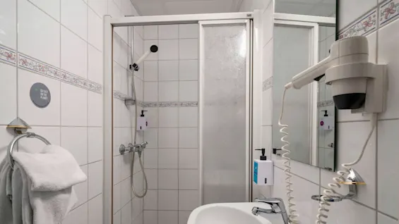 A white-tiled bathroom features a shower with a handheld nozzle, a wall-mounted hairdryer, and a towel on a ring. Text: "GO EASY ON THE PLANET" displayed on a round sign.