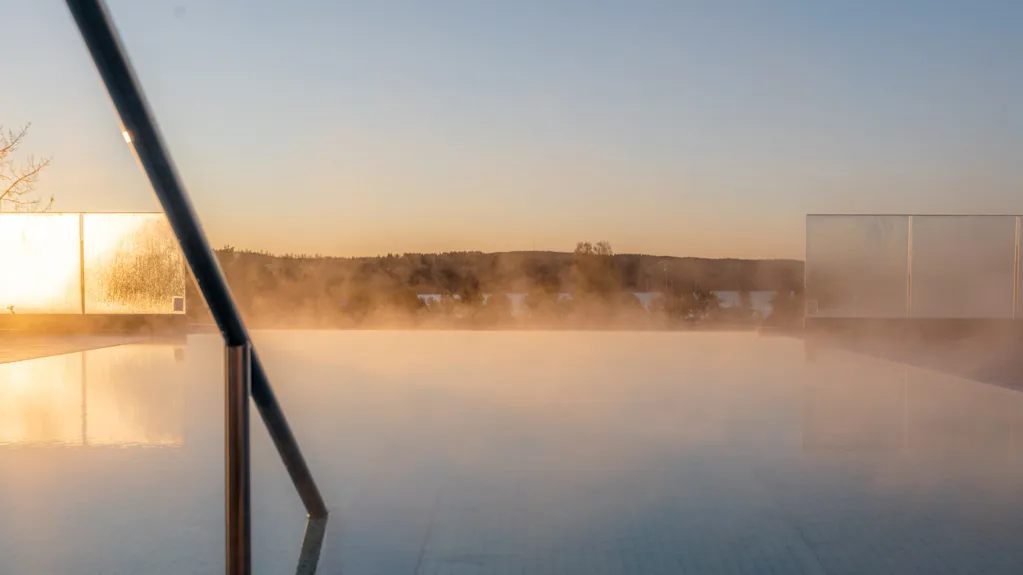 A steaming pool with a metal handrail is situated outdoors during sunset, surrounded by trees and distant hills under a clear sky.