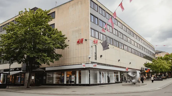 En hjørnebygning huserer en H&M-butikk med et tre foran. Fargede flagg henger over hodet. Folk går langs gaten.