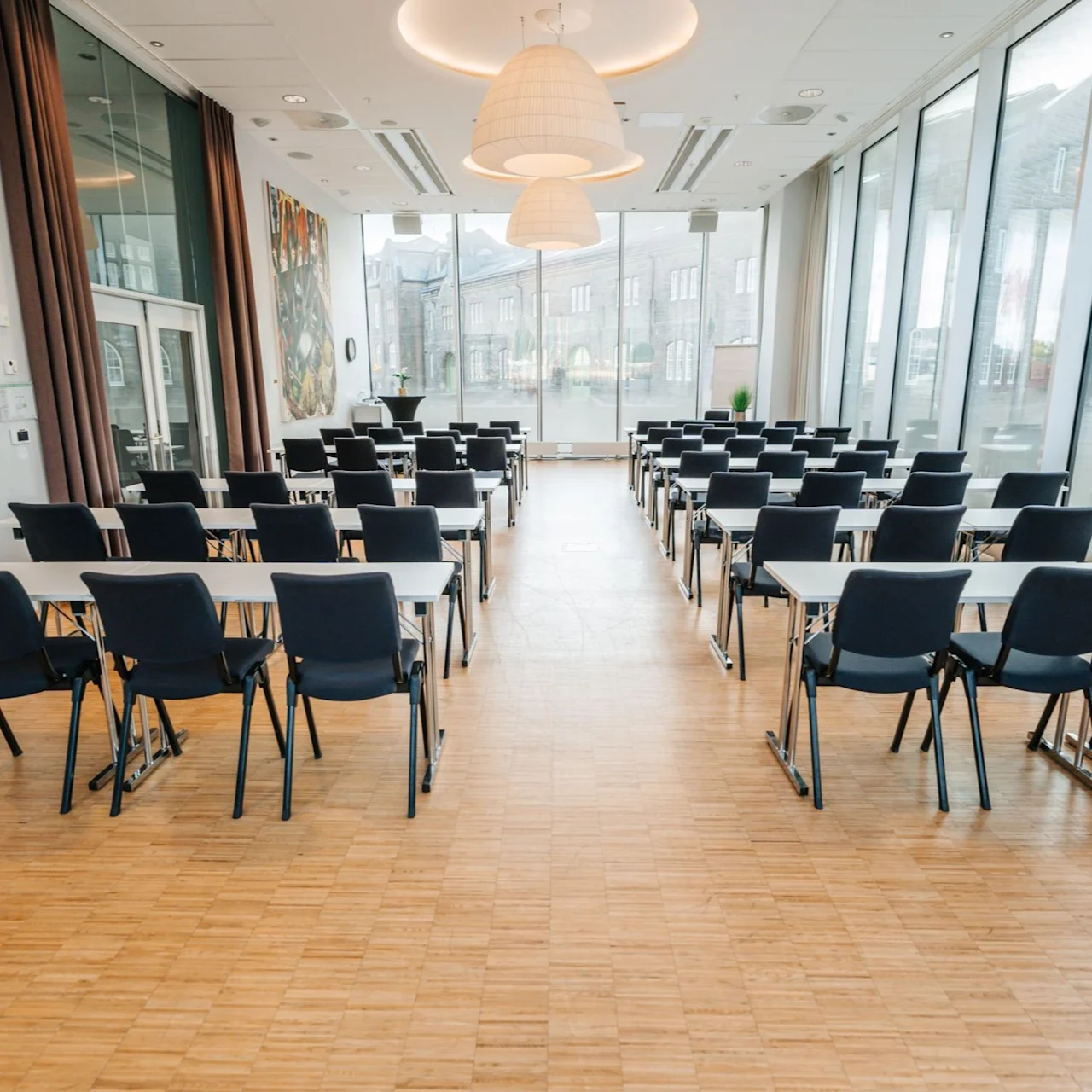 Rows of empty black chairs face forward, arranged with white tables in a spacious, sunlit room. Large windows line the right wall, and vibrant artwork decorates the left side.