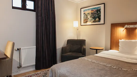 A cozy hotel room at Home Hotel Kung Oscar with a bed, armchair, and a scenic painting on the wall.