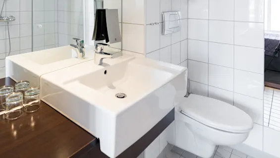Et moderne bad med en rektangulær hvit vask og en kromkran på en trebenk. Fliser på vegger og gulv omgir det, med et speil og håndklestang i sikte.