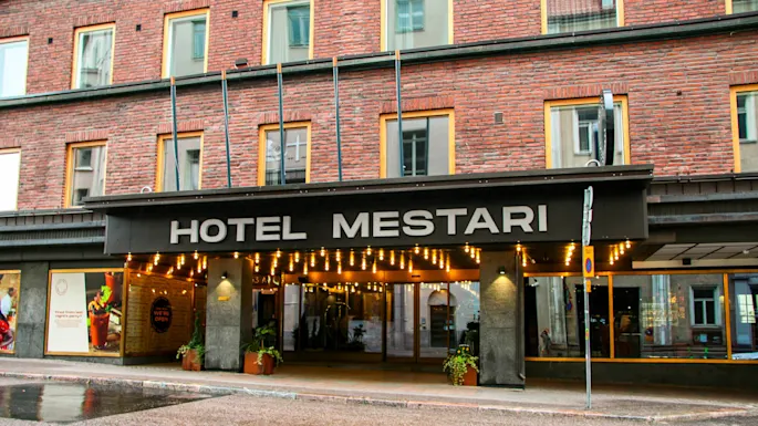 En hotellinngang viser navnet "HOTEL MESTARI" under lysende lys. Murbygningen har store vinduer. Et skilt lyder "WE’RE OPEN" med planter ved inngangen.