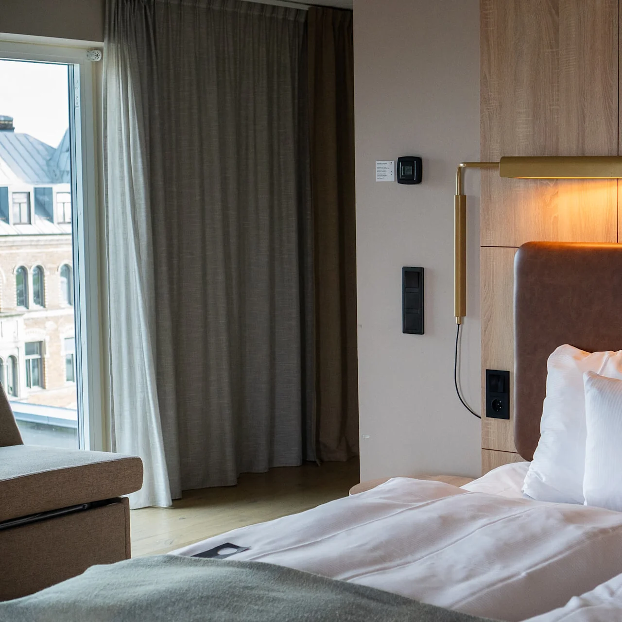 En seng med hvite og mønstrede puter er opplyst av en veggmontert lampe. Den står mot en treplate ved siden av et vindu med gardiner, med utsikt over bygninger utenfor.