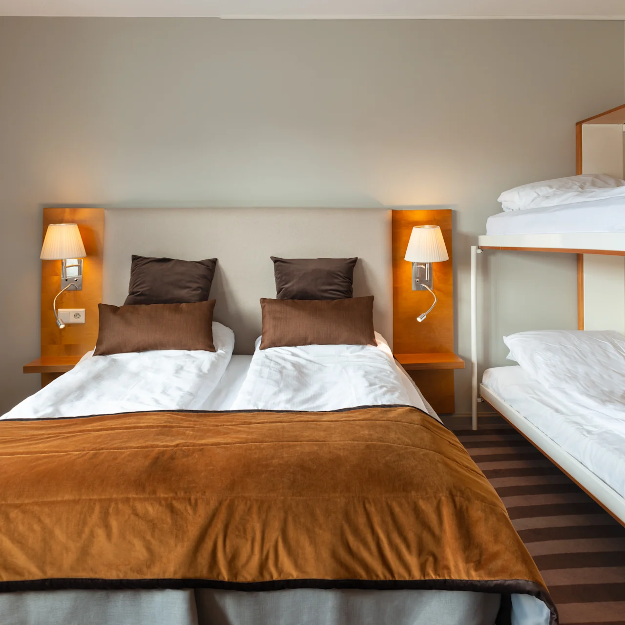 A double bed with brown pillows and covers stands against a wall with two wall lamps. Adjacent, there are white bunk beds in a carpeted hotel room.
