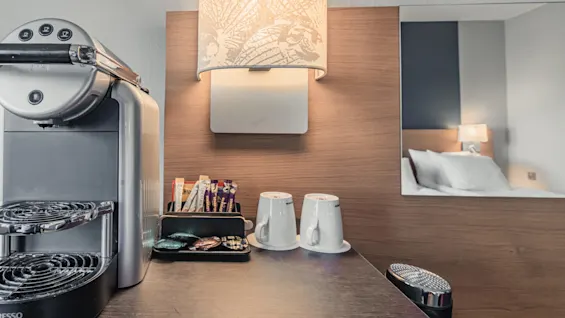 Espresso machine sits idle on a wooden countertop, next to coffee pods, stirrers, and two cups; a wall lamp illuminates the area, while a bed is visible in reflection.