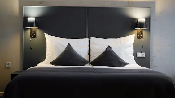 A neatly made double bed with white and dark pillows, flanked by two wall-mounted lamps, in a modern hotel room with concrete walls and a dark headboard.
