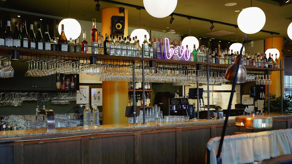 Puinen baaritiski laseineen, viinapulloineen ja neonkyltti, jossa lukee "bar", sisällä lämpimän valaistussa tilassa.
