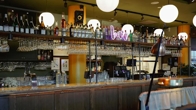 Trebardisk med glasservise, brennevinsflasker og et neonskilt som sier "bar" inni en varmt opplyst etablering.