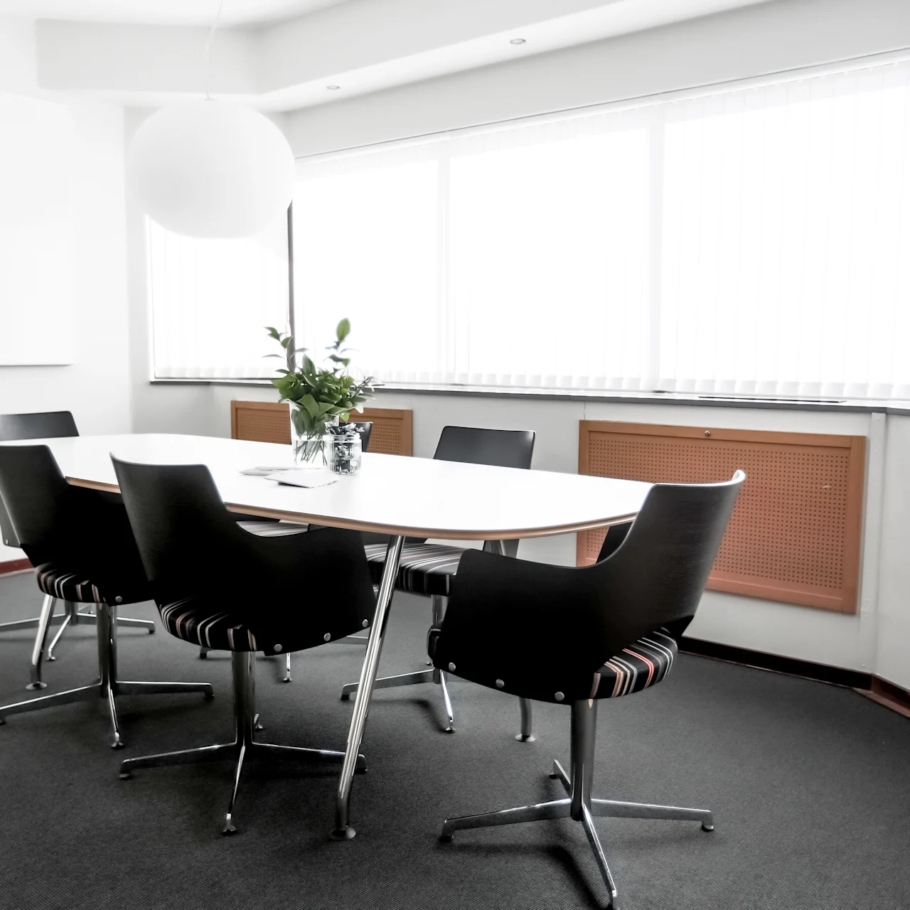 A white conference table surrounded by six black chairs stands in a bright room with large windows, carpeted floor, and a potted plant centerpiece on the table.