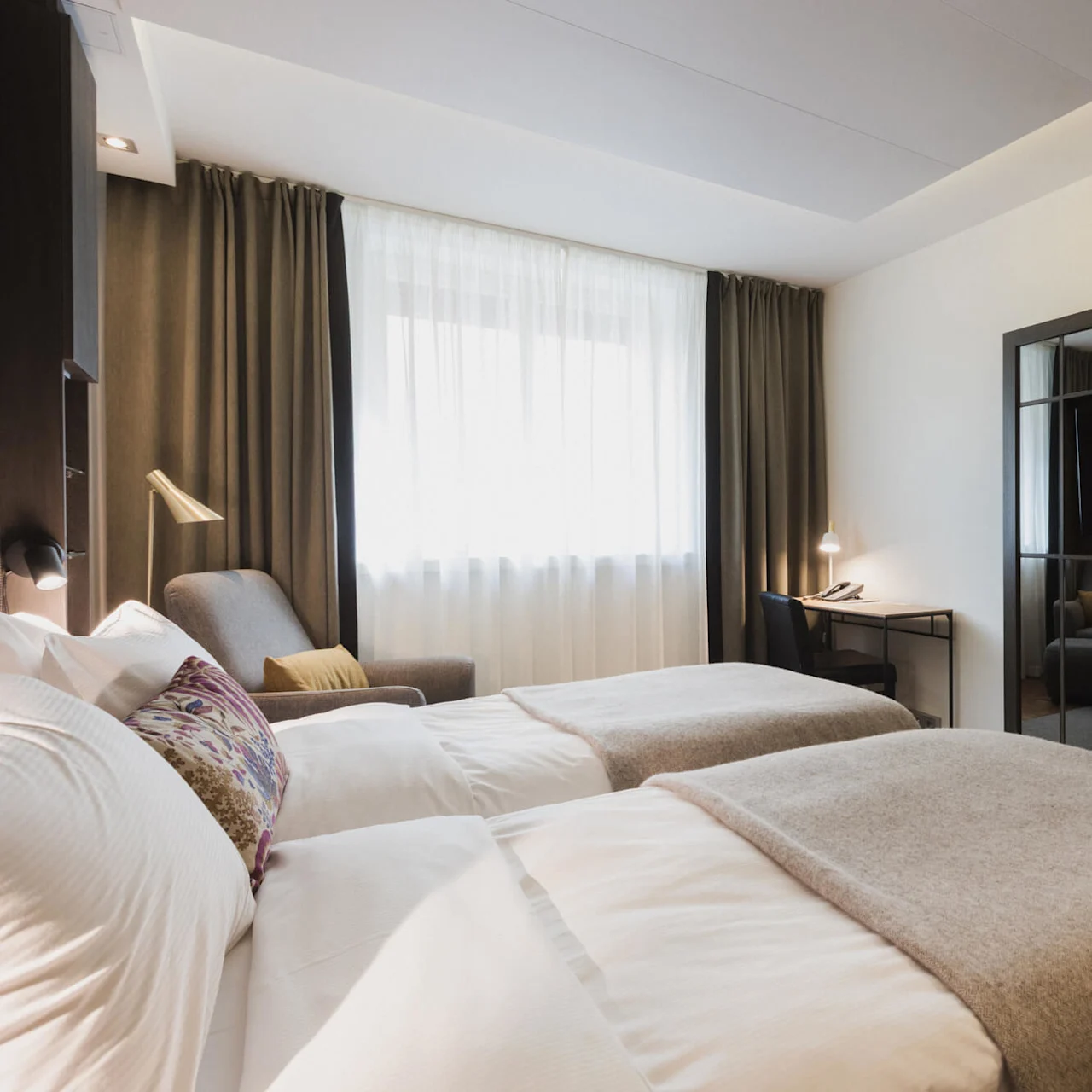 To omhyggeligt redte enkeltsenge står i et moderne hotelværelse, med en grå lænestol og skrivebord foran store draperede vinduer, reflekteret i et vægspejl.