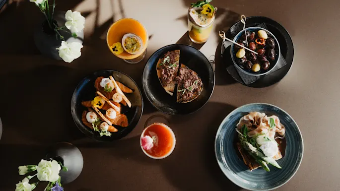 Flere elegante retter er kunstnerisk arrangert på et mørkt bord. Hvert fat inneholder fargerik mat, pyntet med blomster. Drinker følger med, innrammet av myke skygger og blomsterdekorasjoner.