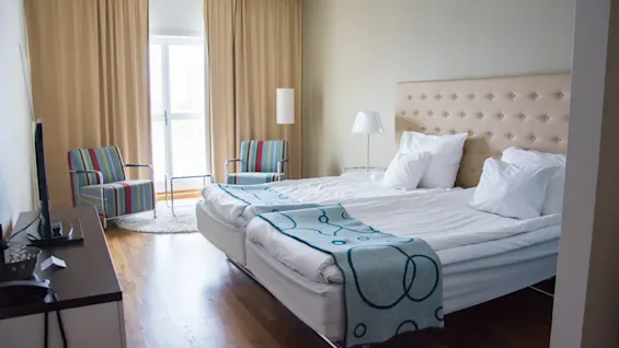 Et lyst og indbydende hotelværelse med to komfortable senge, stribede lænestole, et TV og store vinduer med beige gardiner.