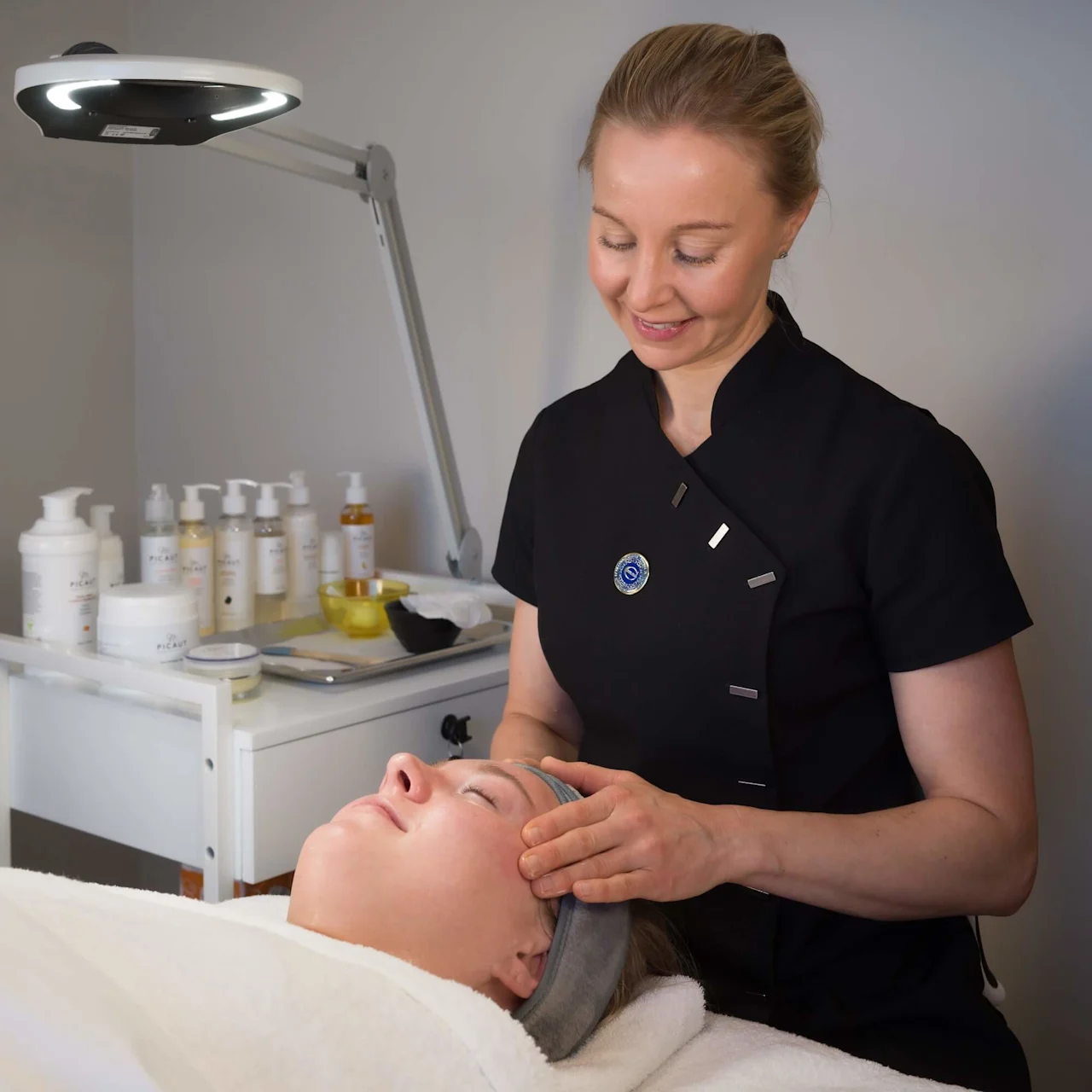 A woman in a black uniform gently massages a client's face under a bright lamp, surrounded by skincare products on a trolley. 