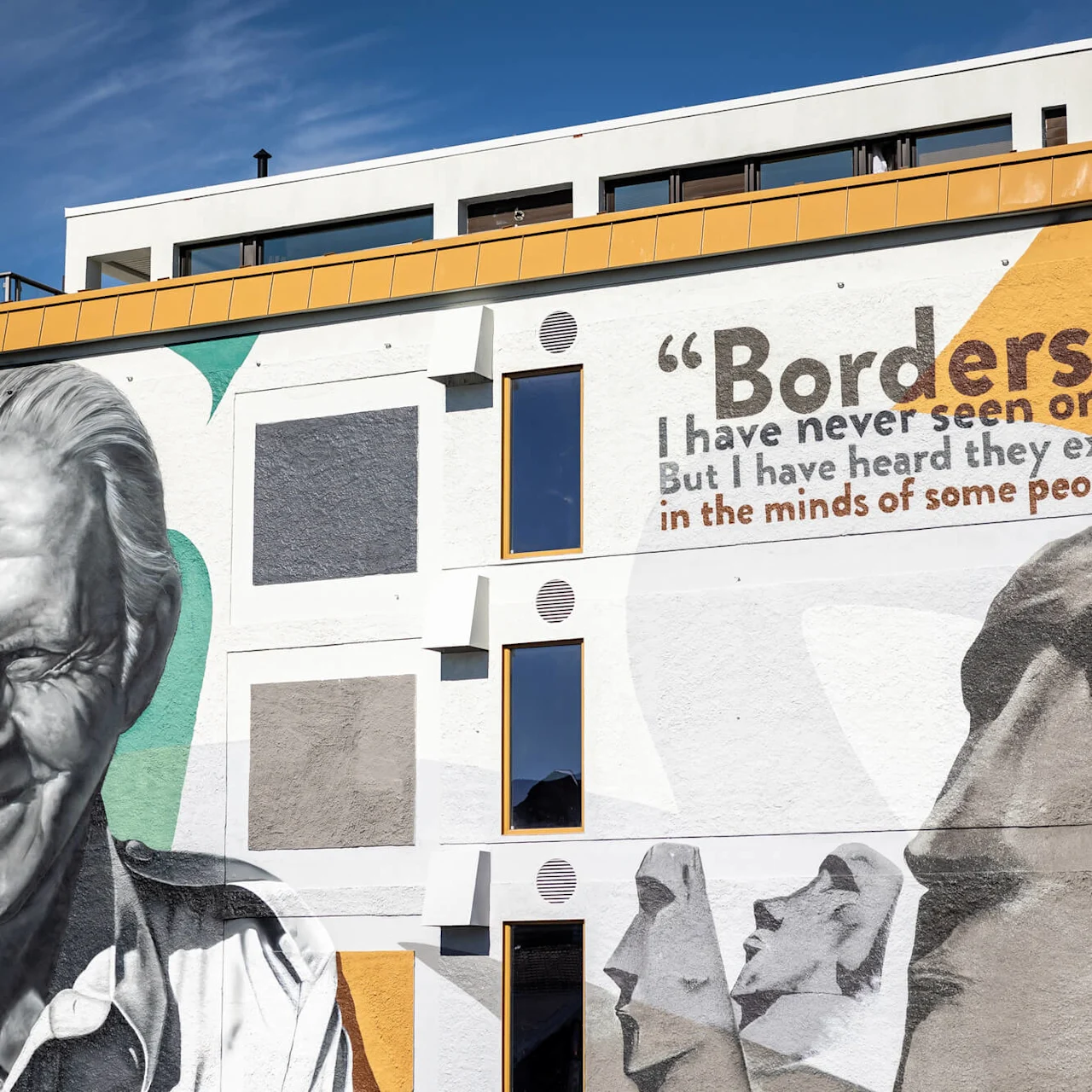 Et farverigt vægmaleri viser en smilende mand og moai-statuer. Teksten lyder: "Borders? I have never seen one. But I have heard they exist in the minds of some people.” Maleriet dækker en bygning i bymiljø.