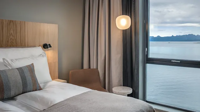 Ett mysigt sovrum med en fint bäddad säng bredvid ett fönster. En stol och ett runt bord står i närheten med utsikt över havet.