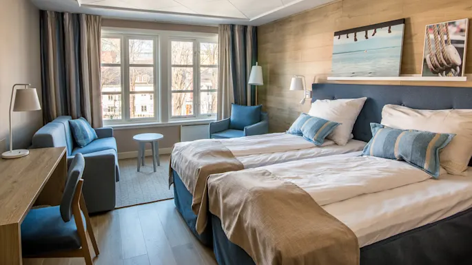 A hotel room with two neatly made beds and a wooden desk; natural light floods through large windows, displaying an urban scene outside.