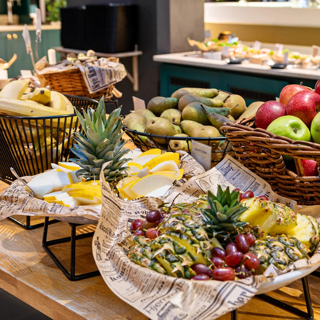 En kurv inneholder røde og grønne epler; i nærheten er det pærer og bananer. Ananas- og melonskiver er kunstnerisk presentert på rustikt papir. Oppsettet er i en bufféomgivelse.