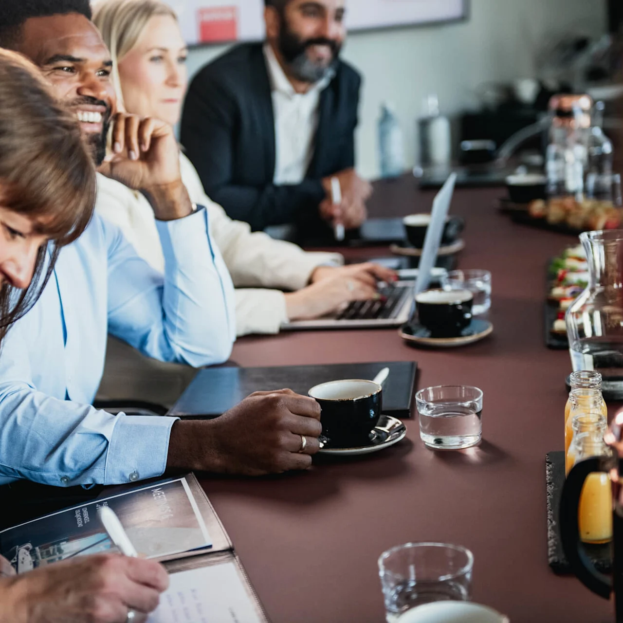 A group of people sits around a table during a meeting. They are jotting notes, using laptops, and drinking coffee. The table is lined with drinks and snacks.