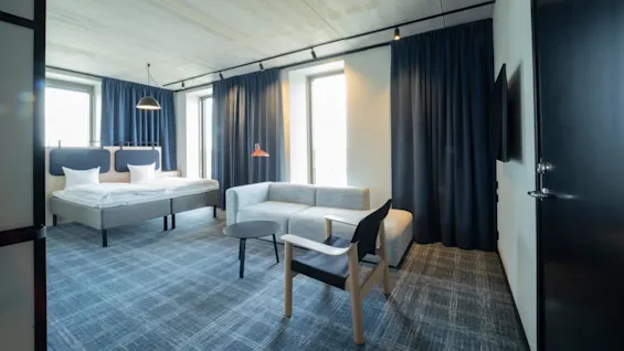Moderni hotellihuoneessa on sänky, sohva ja pieni pöytä hyvin valaistussa tilassa, jossa on siniset verhot ja matto.
