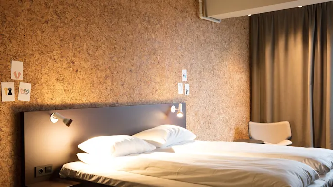 Bed with white sheets under two wall-mounted lights in a room. Decor includes minimalist art on a corkboard wall and a beige chair near a closed curtain.