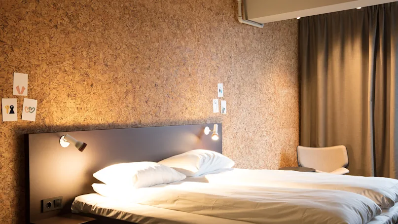 Bed with white sheets under two wall-mounted lights in a room. Decor includes minimalist art on a corkboard wall and a beige chair near a closed curtain.