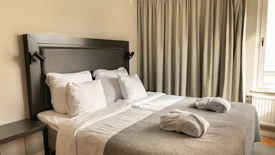 En prydligt bäddad hotellsäng med vita lakan, ett grått överkast och två badrockar, redo för en gästs vistelse.