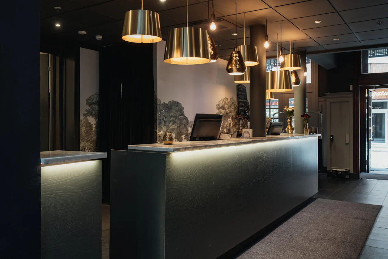 Et indbydende hotelreceptionsområde med en moderne disk med varm belysning, messingpendellamper og et naturinspireret vægmaleri.