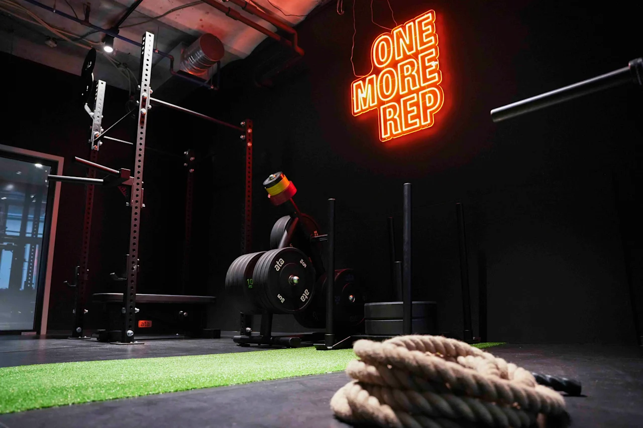Et squat rack og træningsudstyr står ubrugte i et svagt oplyst fitnesscenter. Neonskilt siger "ONE MORE REP". Tyk reb ligger rullet op på grønt græs. Industrielt loft synligt.