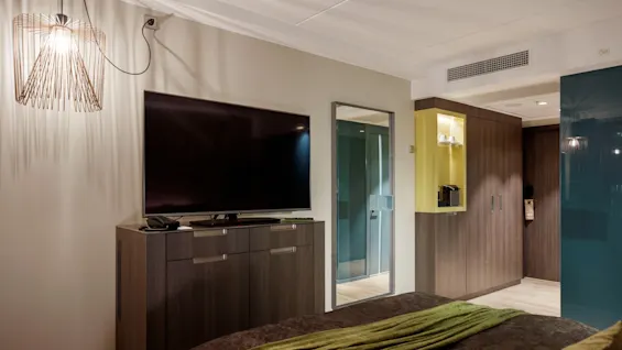 A wall-mounted flat-screen TV is off, above a dark wood cabinet, in a modern bedroom with a bed in foreground. A lamp hangs on the left. 
