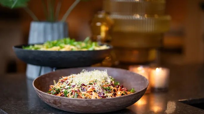 A wooden bowl filled with a mixed salad, featuring shredded carrots and sprouts, sits on a dark countertop. In the background, a second bowl and lit candles provide a warm ambiance.