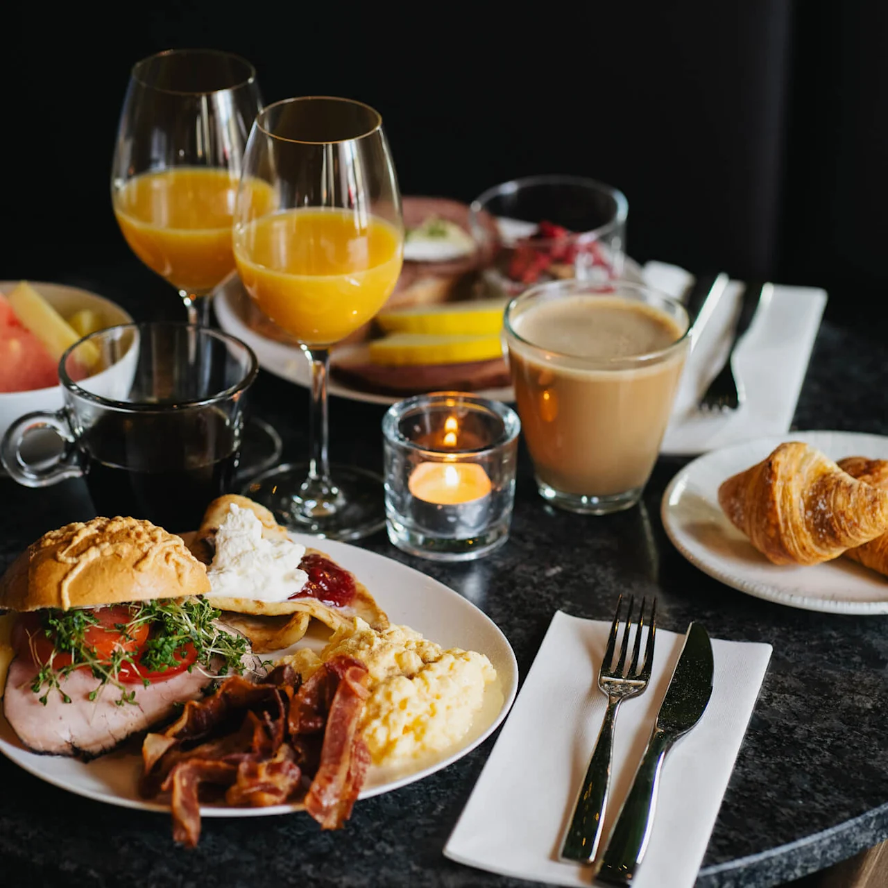 En velpræsenteret morgenmad på et mørkt bord inkluderer croissanter, kaffe, appelsinjuice, bacon, røræg, brød og frugt, med et lys for stemning.