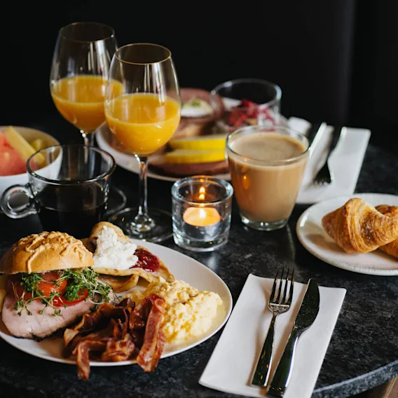 A well-presented breakfast on a dark table includes croissants, coffee, orange juice, bacon, scrambled eggs, bread, and fruit, with a candle for ambiance.