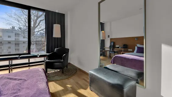 Ett modernt hotellrum med en säng med lila sängkläder och utsikt från ett soligt fönster.