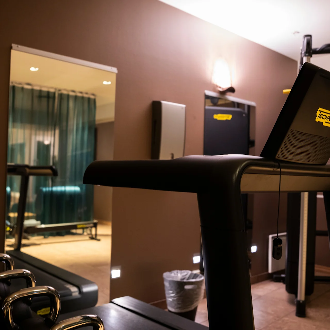 Treadmill positioned centrally, surrounded by dumbbells and a mirror in a dimly lit gym with curtains in another area. Display reads "Technogym."