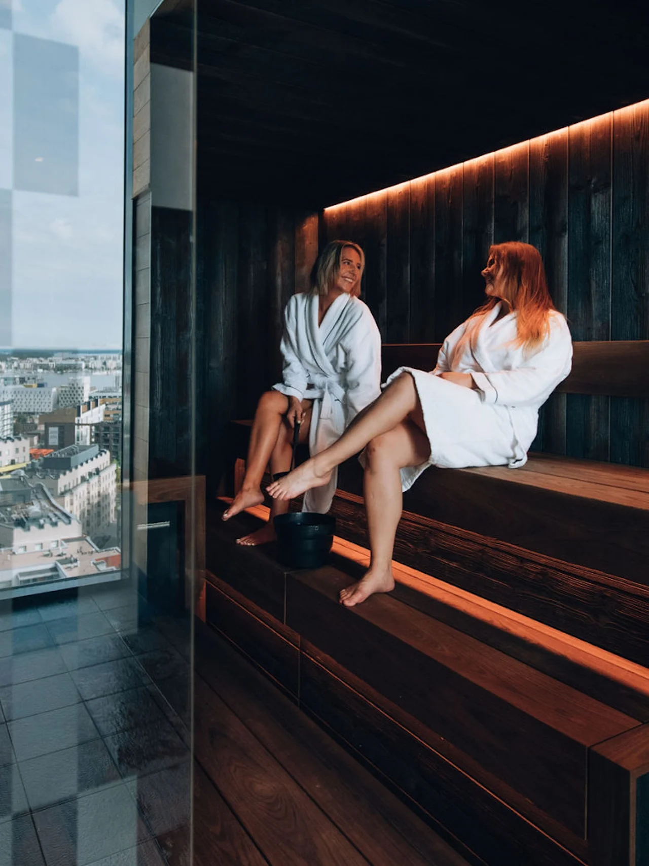 Kaksi kylpytakkiin pukeutunutta henkilöä rentoutuu ja keskustelee modernissa saunassa panoraamanäkymällä kaupunkiin.