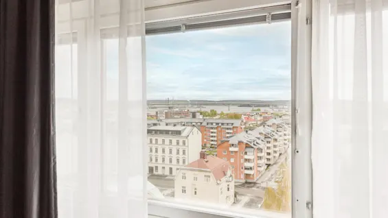 A window view of a city with buildings, a river, and a bridge in the distance, framed by sheer white curtains.