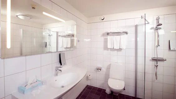 Ett badrum med vita kakelplattor har en handfat, toalett och dusch, samt handdukar på en hylla och modern belysning ovanför.