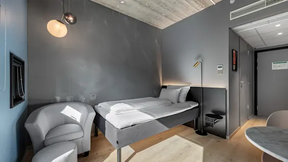 Et moderne hotellrom med enkeltseng, komfortabel lenestol og fotskammel, samt et lite bord med stoler i minimalistisk stil.