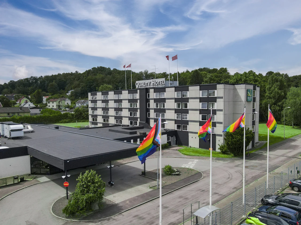 Ett fler våningshotell har regnbågsflaggor vid ingången, omgiven av träd och kullar. Text: "Quality Hotel."